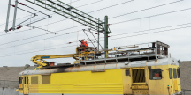 Två arbetare i orangea arbetskläder står uppe på ett gult tåg och arbetar med ledningar.
