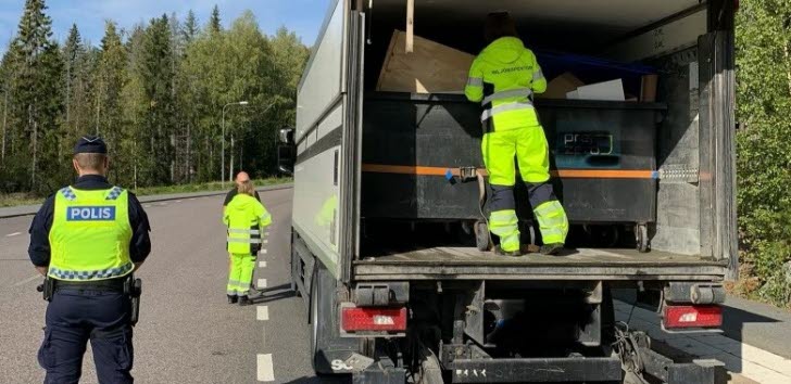 Person i varseldräkt inspekterar lasten på en lastbil. Bredvid bilen, på vägen, står en polis. 