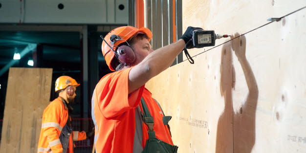 En person i hjälm och med hörselskydd och handskar borrar i en vägg. I bakgrunden står ytterligare en person i skyddskläder.