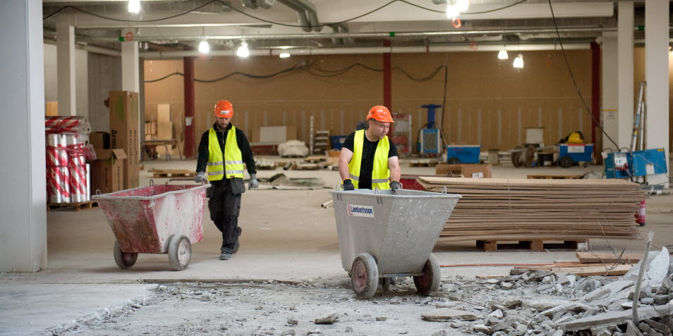 Två byggarbetare går på en byggarbetsplats med varsin portabel rullcontainer framför sig.